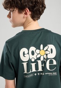 Vihreä puuvillapaita, jossa on suuri valkoinen grafiikka, jossa on keltainen kukka ja teksti "HYVÄ Elämä" takana, sileä pinta, pyöreä kaula-aukko.