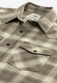 Béžovo-hnědá károvaná košile s černými patentními knoflíky, náprsní kapsou s klopou a límcem, položena na rovné bílé ploše.