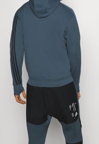 Felpa blu con finitura textured e strisce nere sulle maniche, abbinata a pantaloni corti neri con dettagli grafici bianchi.