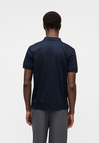 Μπλε ναυτικό πόλο  πουκάμισο με υφασμένη εξαγωνική υφή, κοντά μανίκια και κλασικό γιακά, συνδυασμένο με γκρι παντελόνια. Εμφανίζεται από πίσω.