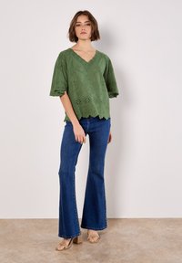 Grünbluse mit gewelltem Rand und Lochdetails, kombiniert mit ausgestellten blauen Jeans und beigen Blockabsatzsandalen, vor einem hellen Hintergrund.