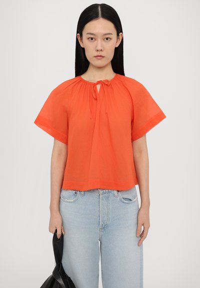 Kurzärmliges, leuchtend orangefarbenes Top mit einem Krawatten-Halsauschnitt und gerafften Details; kombiniert mit hellblauen, hochgeschnittenen Jeans und einer schwarzen Tasche.