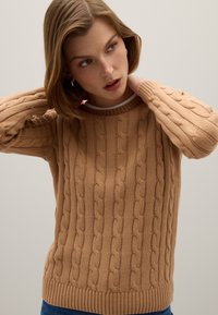 Suéter de punto cableado en marrón claro con un cuello y puños acanalados, que presenta un patrón texturizado y un corte holgado. Material de mezcla de lana.