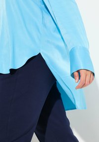 Tunika azzurra chiara in seta con ampi polsini, orlo ondulato e spacco laterale, abbinata a pantaloni blu scuro. Texture morbida e design fluido.