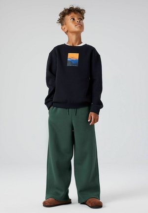 Zwart sweatshirt met een kleurrijke bergafbeelding; gecombineerd met losse groene broek en bruine slippers, met boorden en een trekkoord.