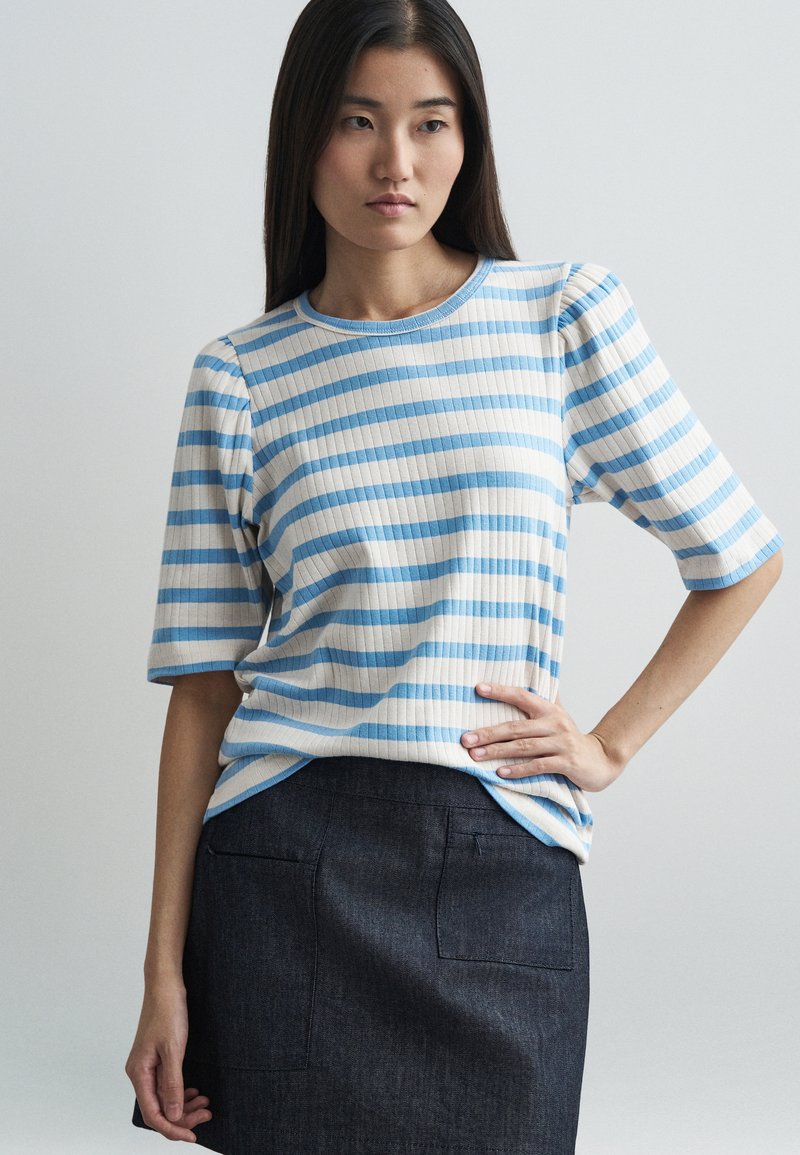 Απαλή μπλούζα με ριγέ σχέδιο σε ανοιχτό μπλε και �κρεμ, με στρογγυλή λαιμόκοψη και κοντά μανίκια, συνδυασμένη με μια σκούρα τζιν φούστα.