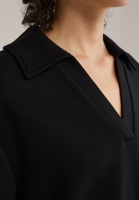 Fekete pulóver V-nyakkal és hajtott gallérral. Puha anyagból készült, sima textúrájú és minimális részletezéssel.