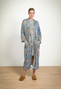 Woman standing on wood floor wearing a blue floral robe and dress with brown slippers, hands in robe pockets, neutral background.