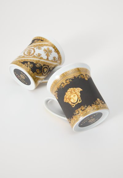 Δύο πορσελάνινα φλυτζάνια με περίπλοκα χρυσά σχέδια. Το ένα έχει μαύρη βάση με κεντρικό μοτίβο προσώπου· το άλλο είναι λευκό με χρυσές λεπτομέρειες.