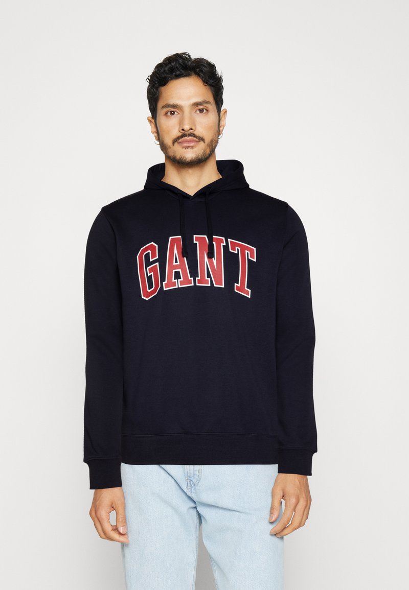 Felpa blu navy con il logo "GANT" in rosso ben visibile, cappuccio con coulisse e polsini a costine. Realizzata in tessuto morbido, ideale per un abbigliamento casual.