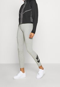 Černá bunda na zip s geometrickým vzorem, doplněná šedými legínami s černým logem Nike a bílými teniskami.