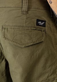 Olivgrüne Shorts aus strukturiertem Stoff, mit einer Gesäßtasche und einem schwarzen Etikett mit weißem Text, das aufgenäht ist.
