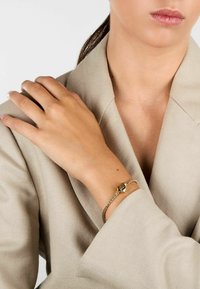 Bracciale d'oro con un charm a forma di cuore sul polso, caratterizzato da un design intrecciato. Il modello indossa un blazer beige chiaro. Texture liscia.