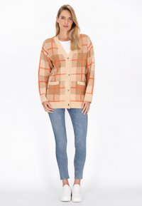Cardigan beige con un motivo a quadri arancione e marrone, dotato di scollatura a V, chiusura con bottoni, due tasche e polsini a costine. Abbinato a jeans blu.