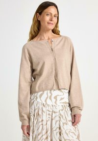 Krémový zkrácený cardigan s kulatým výstřihem a zapínáním na knoflíky, doplněný bílou sukní s krémovým zebrovaným vzorem. Měkká textura.