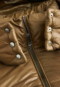 Brun pufferjakke i stof med skinnende tekstur, med lynlås foran og trykknapper på kraven. Kvalitetens quiltede design er synligt.