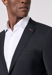 Completo nero su misura con camicia bianca. Caratterizzato da una vestibilità slim, texture liscia e un piccolo emblema rosso sulla patta.