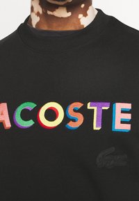 Svart sweatshirt med färgglada broderade bokstäver som stavar "LACOSTE", med en texturerad logotyp i det nedre hörnet.