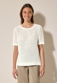 Bílý pletený top s krátkým rukávem, kulatým výstřihem a texturovaným otevřeným vzorem. V kombinaci s béžovými, volně střiženými kalhotami.