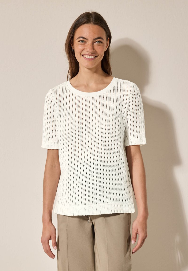 Bílý pletený top s krátkým rukávem, kulatým výstřihem a texturovaným otevřeným vzorem. V kombinaci s béžovými, volně střiženými kalhotami.