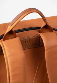 Brauner Stoff-Rucksack mit glatter Oberfläche, ausgestattet mit einem Ledergriff, gepolsterten Trägern und einem Logo-Patch. Stilvolles und funktionales Design.