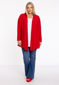 Rode lange mouwen cardigan met een open voorkant, gecombineerd met een witte top en een blauwe flared jeans. Het model draagt rode plateau schoenen.