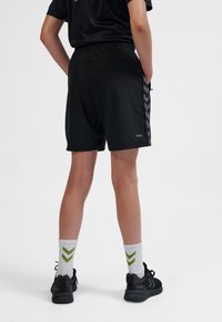 Pantaloncini sportivi neri con vita elastica, caratterizzati da un pannello laterale a motivo e un logo sulla parte anteriore inferiore. Abbinati a scarpe nere e calzini con strisce verdi.