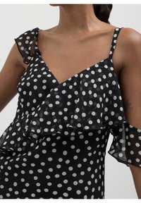 Robe noire à pois avec bretelles à volants, dotée d'un décolleté en V et de volants superposés. Le tissu semble léger et texturé.