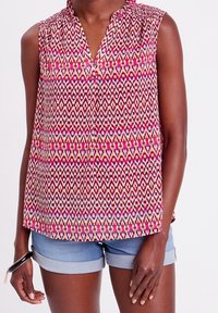 Haut sans manches rose avec un motif géométrique en rouge, blanc et vert. Col classique, tissu léger, design à longueur de hanches.