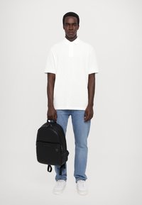 Polo shirt blanc texturé, jeans bleu clair, sac à dos noir avec fermeture éclair, et baskets blanches. Le modèle se tient avec les bras le long du corps.