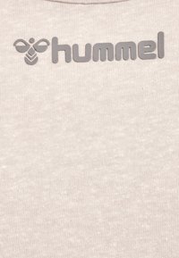 Das Logo der Marke Hummel zeigt ein stilisiertes Bienen-Icon und Kleinbuchstaben auf einem hellbeigen, strukturierten Stoffhintergrund.
