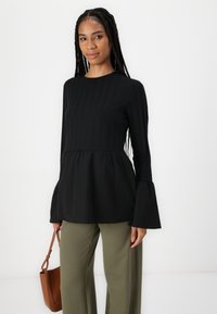 Top peplum nero con maniche lunghe a campana e texture a coste verticali; abbinato a pantaloni a gamba larga verde oliva e una borsa marrone.