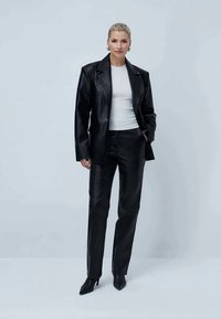 Woman with blonde hair wearing black leather blazer and pants, white fitted top, black heeled boots, standing with one hand in pocket.