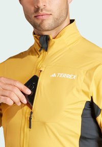 Yellow Terrex jacket with black accents, high collar, and zippered pocket; smooth texture, streamlined design, suitable for outdoor use.