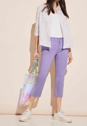 Pantalones morados recortados, camiseta blanca y una camisa de rayas en lavanda. Sosteniendo un bolso tote iridiscente. Zapatillas blancas con suelas texturizadas.
