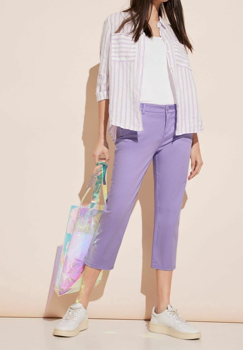 Fioletowe spodnie do połowy łydki, biała koszulka i pasiasta lawendowa koszula. Trzyma perłową torbę na ramię. Białe trampki z teksturowaną podeszwą.