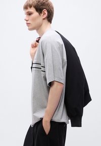 Γκρι κοντομάνικο T-shirt με μαύρες ρίγες, φορεμένο με μαύρο σακάκι περασμένο πάνω από τον έναν ώμο, συνδυασμένο με μαύρο παντελόνι.