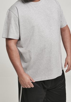 Man draagt een lichtgrijs T-shirt met korte mouwen en zwarte sportshorts met witte strepen, staat met ontspannen handen langs het lichaam.