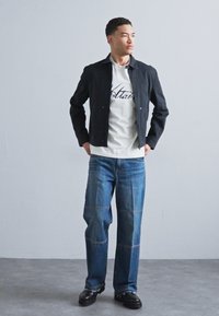 Black cropped jacket over a white shirt with graphic text, paired with blue jeans featuring contrasting seams and black chunky shoes.