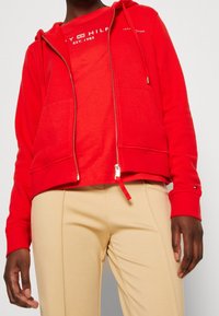 Sweat à capuche rouge zippé avec une poche kangourou, des poignets côtelés et un logo imprimé. Porté par-dessus un t-shirt rouge, accompagné d'un pantalon beige.