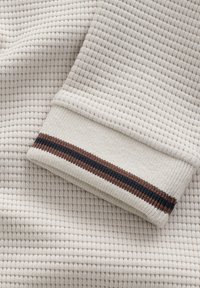 Ljust beige strukturerad tyg med ett våffelmönster, med en mudd med en krämfärgad bas och en brunt och marinblå randig detalj.