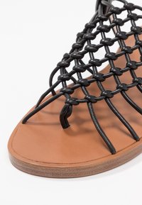 Sandales à tige tissée noire avec une semelle en caoutchouc beige. Elles présentent des brides fines et un design ouvert à l'avant, mettant en avant une structure minimaliste et respirante.