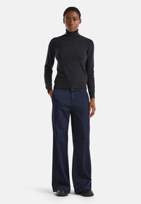 Femme debout, portant un pull à col roulé gris foncé, un jean large bleu foncé et des chaussures noires, les mains dans les poches, sur un fond neutre.