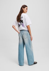 Maglietta bianca corta con un design grafico sulla schiena, abbinata a jeans larghi di un blu chiaro scoloriti e scarpe slip-on nere con borchie.
