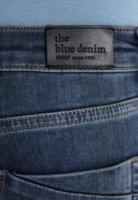 Jeans in denim blu scuro con superficie testurizzata. Caratterizzati da un'etichetta in pelle nera con testo in rilievo che recita "the blue denim ONLY Anno 1995."