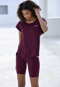 Burgundijas aktīvās apģērba komplekts ietver īsām piedurknēm topu un piegulošas šortus, ar gludu faktūru un zīmola nosaukumu baltā krāsā abās daļās.