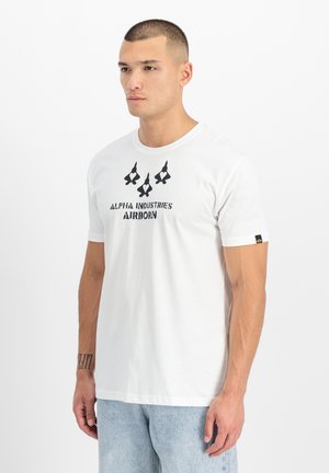 Man wearing white T-shirt with black "Alpha Industries Airborn" text and three jet silhouettes with heart symbols above light blue jeans.