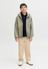 Garçon debout portant une veste beige sur un polo bleu marine, un pantalon beige et des baskets blanches sur un fond clair uni.