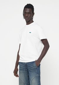 Camiseta blanca de algodón con cuello redondo, que presenta un pequeño logotipo de cocodrilo bordado en verde en el lado izquierdo del pecho. Se lleva con pantalones cortos de mezclilla azul.