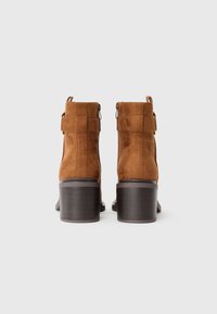 Botas de tobillo de ante marrón con tacón cuadrado y cremalleras en la parte trasera, con un diseño minimalista y detalles de costura vertical.